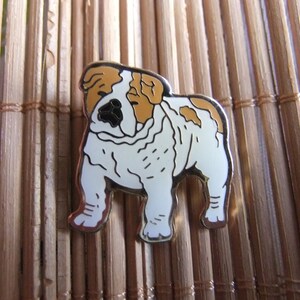 Könnte beinhalten: Metall-Anstecknadel mit einem Bulldoggen-Motiv. Die Anstecknadel zeigt einen weißen Bulldoggen mit braunen Flecken und schwarzen Details, umrandet von Silber. Der Bulldogge ist in einer stehenden Position dargestellt, mit deutlich definierten Merkmalen.