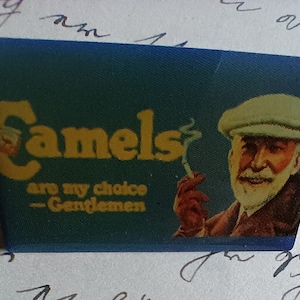 Op de afbeelding: Een blauw en geel vintage reclamebord voor Camel-sigaretten. Het bord toont een man met een witte pet die een sigaret rookt en de tekst "Camels are my choice - Gentlemen".