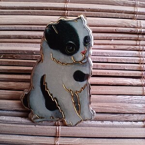 May include: A black and white enamel pin of a cat sitting with its head tilted. The cat has a black nose and a pink mouth.
