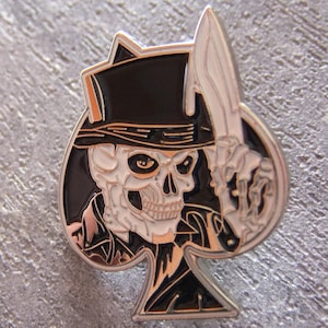 Op de afbeelding: Een zilveren metalen pin in de vorm van een schoppen speelkaart met een schedel die een zwarte hoge hoed draagt en een mes vasthoudt. De schedel heeft witte ogen en een wit gezicht.