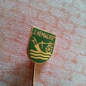 Könnte beinhalten: Eine Vintage-Anstecknadel mit grünem Schilddesign. Das Schild zeigt das Wort "EHEMALIGE" oben, mit einer Schaufel, Wasser und einem Raddesign. Die Nadel hat einen goldfarbenen Metallstiel.