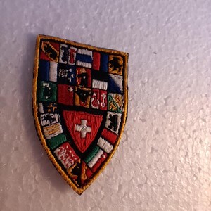 Könnte beinhalten: Ein farbenfroher gestickter Aufnäher in Form eines Schildes mit einer Schweizer Flagge in der Mitte. Der Schild ist von kleineren Flaggen verschiedener Länder umgeben.