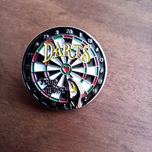 Puede incluir: Un tablero de dardos blanco y negro con secciones rojas y verdes. El tablero de dardos tiene la palabra "DARTS" escrita en letras doradas en el centro. Una mano sostiene un dardo, apuntando al centro del tablero, que es un corazón rojo.