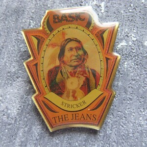 Könnte beinhalten: Eine goldfarbene Metallstifte mit einem roten, orangefarbenen und schwarzen Design. Die Stifte zeigt ein Porträt eines indianischen Mannes mit einem Federkopfschmuck. Der Text "BASIC" befindet sich oben auf der Stifte, und der Text "STRICKER THE JEANS" befindet sich unten.