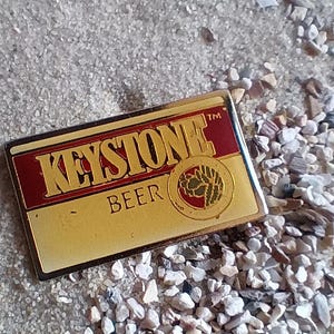 May include: A gold and red metal pin with the words "Keystone Beer" and a logo of a stylized tree. The pin is on a background of small white and brown rocks.
