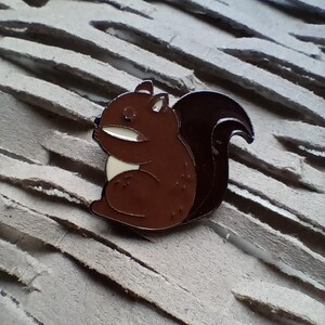 May include: A brown and dark brown enamel pin in the shape of a squirrel. The squirrel is in a sitting position with a white belly and a fluffy tail. The pin has a silver outline.
