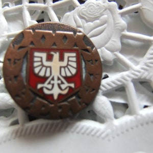 May include: A bronze-coloured pin with a red, white and black shield featuring a white eagle with a cross and anchor. The shield is surrounded by a circle of text that reads "AWW 1939".