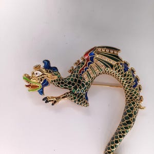 Könnte beinhalten: Goldfarbene Drachenbrosche mit grünen, blauen, roten und weißen Emaille-Details. Der Drachenkörper ist geschwungen, mit einem detaillierten Schuppenmuster. Die Brosche ist mit kleinen, klaren Kristallen verziert.
