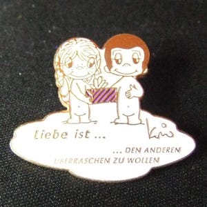 May include: A white enamel pin with a cartoon couple, a woman with blonde hair and a man with brown hair, holding a purple and white striped gift box. The text "Liebe ist... ...den anderen ueberraschen zu wollen" is written on the pin.