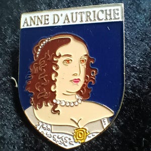 Könnte beinhalten: Schildförmige Emaille-Anstecknadel mit einem Porträt einer Frau mit braunem, lockigem Haar, die eine Perlenkette und ein weißes Kleid trägt. Die Anstecknadel hat einen blauen Hintergrund und den Text "ANNE D'AUTRICHE" oben.