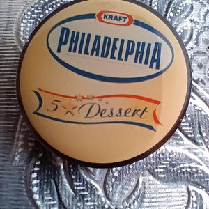 Könnte beinhalten: Ein rundes, cremefarbenes Etikett mit blauem und rotem Rand. Das Etikett trägt die Aufschrift "KRAFT PHILADELPHIA 5-X Dessert" in weißer Schrift.