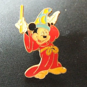 May include: A gold-coloured pin featuring Mickey Mouse dressed as a wizard. He is wearing a red robe with a yellow sash, a blue hat with yellow stars, and is holding a gold wand.