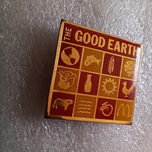 Könnte beinhalten: Eine quadratische Anstecknadel in Gold und Rot mit den Worten "The Good Earth" und einem Raster mit 12 kleinen Bildern, die Natur und Lebensmittel darstellen. Die Bilder zeigen einen Globus, einen Fisch, Weizen, einen Baum, Obst, eine Flasche, die Sonne, ein Huhn, eine Kuh, Wellen, einen Apfel und das McDonald's-Logo.