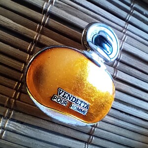 Könnte beinhalten: Eine silberne und goldfarbene Parfümflasche mit dem Text "VENDETTA POUR HOMME" auf der Vorderseite.