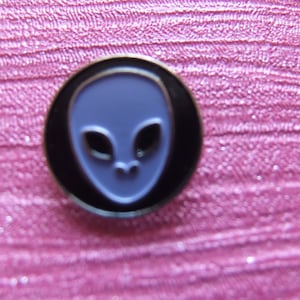 Könnte beinhalten: Eine schwarze und silberne Anstecknadel mit einem lila Alien-Gesichtsdesign.