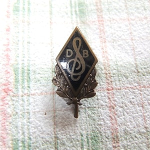 Könnte beinhalten: Eine schwarze und goldene Metallnadel mit einem Violinschlüssel und den Buchstaben "DB" in einer Diamantenform. Die Nadel hat unten ein Blattdesign.