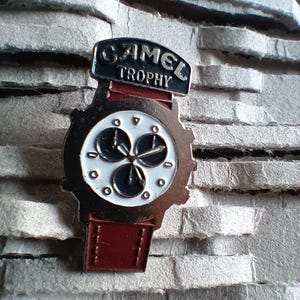 Op de afbeelding: Een vintage Camel Trophy horlogepin met een zilverkleurige wijzerplaat en een rode band. De pin toont de woorden "CAMEL TROPHY" in zwarte letters op een zilveren achtergrond.