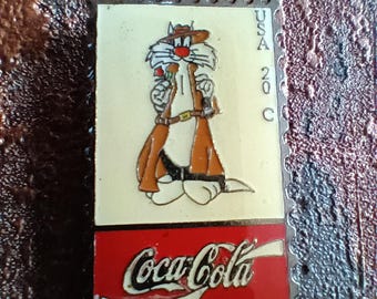 May include: A vintage Coca-Cola collectible pin featuring a cartoon cat wearing a hat and holding a rifle. The pin is shaped like a postage stamp with the text "USA 20 C" and "Coca-Cola" printed on it.