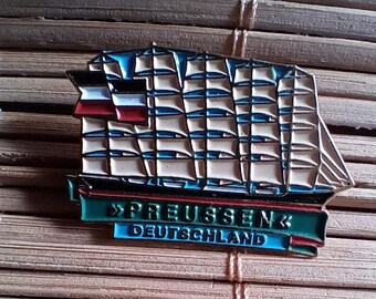 May include: A blue and white enamel pin featuring a ship with a sail and the text "Preussen Deutschland".