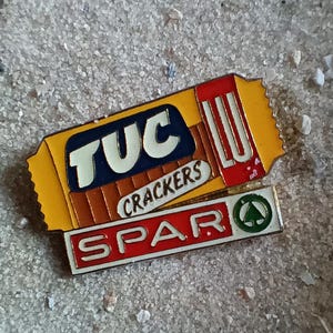 Könnte beinhalten: Eine gelbe und rote Anstecknadel mit dem Text "TUC CRACKERS" und "LU" auf gelbem und braunem Hintergrund. Die Anstecknadel hat auch den Text "SPAR" in Rot und Weiß auf weißem Hintergrund.