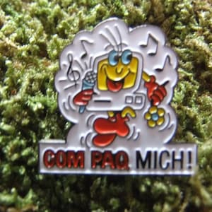 May include: A white enamel pin with a cartoon computer character singing into a microphone. The character is wearing red shoes and has a yellow face. The text "COM PAQ MICH!" is written on a white banner below the character.