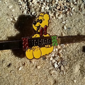 May include: A gold-colored metal pin with a yellow gummy bear holding a red and white gummy bear bar with the word "HARIBO" printed on it. The gummy bear is wearing sunglasses and a red bow tie.