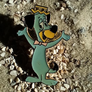 Könnte beinhalten: Ein Vintage-Emaille-Pin mit einer Cartoon-Hundefigur in Helltürkis. Der Hund hat schwarze Ohren, eine gelbe Schnauze und trägt eine goldene Krone und eine rosa Fliege. Der Pin ist vor einem Hintergrund aus kleinen Kieselsteinen platziert.