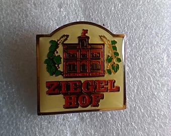 May include: A gold-colored pin with a white background and red lettering. The pin features a red building with a gold roof and a green vine on either side. The text on the pin reads "Ziegel Hof".