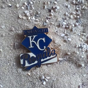 Puede incluir: Un pin de los Kansas City Royals azul y dorado con el logotipo del equipo y las palabras "Kansas City" y "Royals".