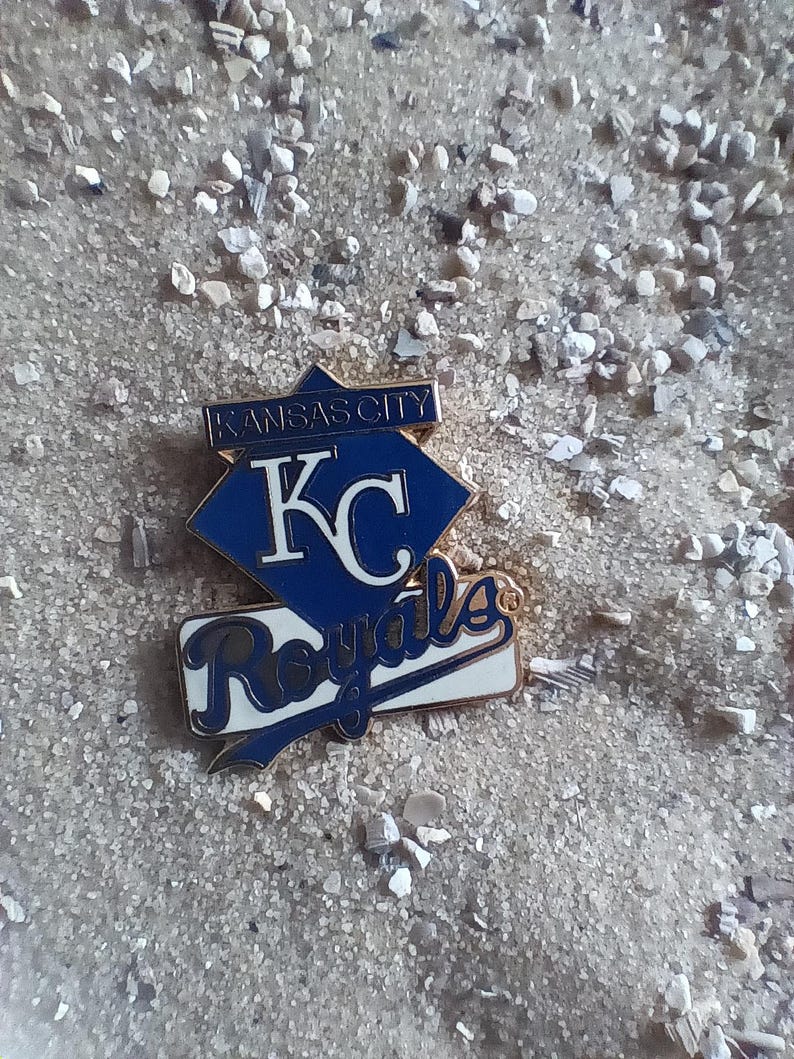Puede incluir: Un pin de los Kansas City Royals azul y dorado con el logotipo del equipo y las palabras "Kansas City" y "Royals".