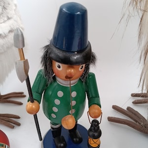 Può includere: Statuina in legno di un soldato con giacca verde, cappello blu e stivali neri. La statuina tiene in mano una lanterna e una lancia. Il soldato è su una base blu. La statuina è un ornamento natalizio.