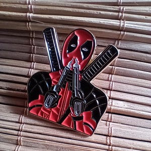 May include: A red and black enamel pin featuring Deadpool holding two guns. The pin is a detailed depiction of the popular Marvel Comics character.