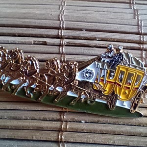 May include: A gold-tone metal pin depicting a horse-drawn carriage with four horses pulling a yellow carriage with a white roof. The carriage has a driver and passengers inside. The pin is detailed and intricate.