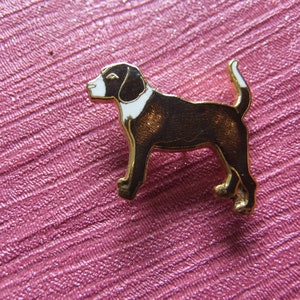 Könnte beinhalten: Eine goldfarbene Metallnadel in Form eines Hundes. Der Hund hat einen braunen Körper, weiße Markierungen auf Gesicht und Brust und eine weißspitzige Rute. Die Nadel befindet sich auf einem strukturierten rosa Hintergrund.