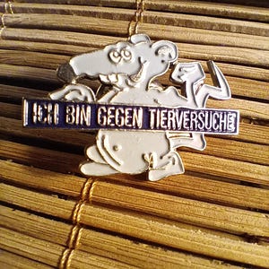 May include: A white and silver pin featuring a cartoon rat holding a hammer. The pin has the text "ICH BIN GEGEN TIERVERSUCHE" in blue letters on a blue background. The pin is set against a bamboo background.