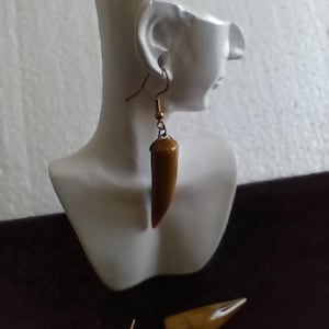 May include: A pair of earrings with a brown, elongated, bullet-shaped design. The earrings are displayed on a white bust and have gold-colored ear wires. One earring is hanging from the bust, and the other is lying on a dark surface.