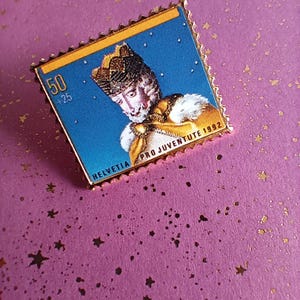 Könnte beinhalten: Quadratischer Anstecker, der einer Briefmarke ähnelt, mit einem Porträt einer gekrönten Person vor blauem Hintergrund. Mit goldfarbenen Rändern und dem Text "HELVETIA PRO JUVENTUTE 1992".