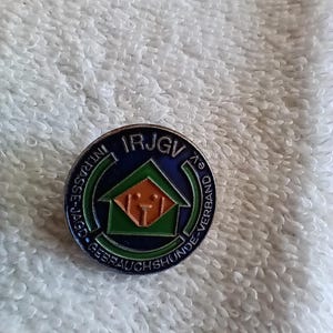 May include: A silver pin with a blue, green, and orange design. The pin features a house with a dog inside and the text "IRJGV" and "Gebrauchshunde-Verband".