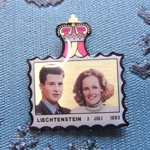 Könnte beinhalten: Eine goldfarbene Anstecknadel mit einer königlichen Krone oben und einem Bild eines Paares in der Mitte. Die Anstecknadel hat einen weißen Rand und den Text "LIECHTENSTEIN 3 JULI 1993" unten.