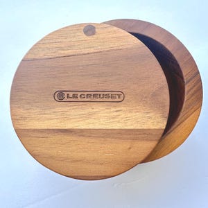 以下が含まれることがあります： 円形の木製容器と、それに対応する円形の蓋。蓋には木目がはっきりと見え、中央にはLE CREUSETというブランド名が刻印されています。容器は開いた状態で、収納用の空洞の内部空間が見えています。