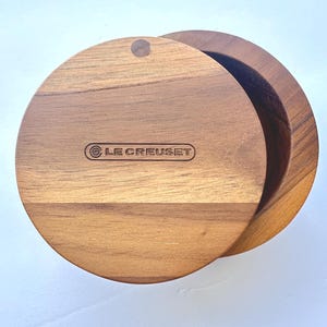 May include: A wooden Le Creuset round lid and serving dish. The lid has a small hole and the Le Creuset logo is visible. The dish is made of wood and has a natural finish.