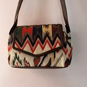 May include: A shoulder bag with a colorful geometric pattern in red, black, white, and green. The bag has a brown leather flap and strap. The bag is rectangular and has a flap closure.