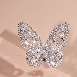 May include: A silver butterfly ring with clear crystal accents. The ring is set against a light pink background.