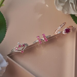 May include: Three rings featuring vibrant red stones. One ring is shaped like a crown, another has two parallel vertical bars set with red stones, and the third is a rose gold-toned band with a central oval red stone and small clear accents.