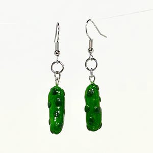 Puede incluir: Un par de pendientes novedosos con dijes de pepinillos verdes en miniatura con protuberancias de color verde oscuro. Los pendientes tienen alambres para la oreja de gancho de pez de color plateado y pequeños anillos de conexión.