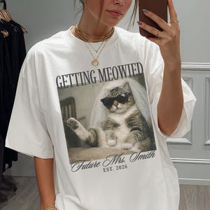 以下が含まれることがあります： サングラスとベールをつけた猫のグラフィックが特徴の白いオーバーサイズのTシャツ。「GETTING MEOWIED」の文字が画像の上にあり、「Future Mrs. Smith EST. 2026」が下にあります。Tシャツは人が着ています。