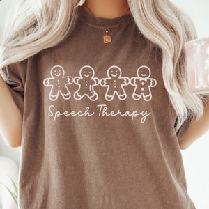 May include: A brown t-shirt with a white graphic of five gingerbread people holding hands and the text "Speech Therapy".