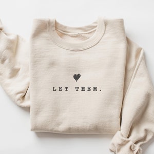 May include: A beige sweatshirt with the text "LET THEM." and a small black heart above the text.