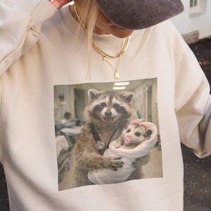May include: A beige crewneck sweatshirt featuring a graphic print of a raccoon dressed as a doctor with a stethoscope, holding a small opossum wrapped in a white blanket with a pink pacifier. The sweatshirt has long sleeves and a ribbed neckline.