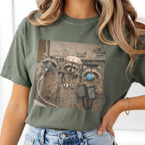 May include: Olive green t-shirt featuring a graphic of three raccoons in medical attire. One raccoon wears a nurse's cap, another has a blue bubble on its nose, and the third holds medical equipment. The image is a humorous take on healthcare.